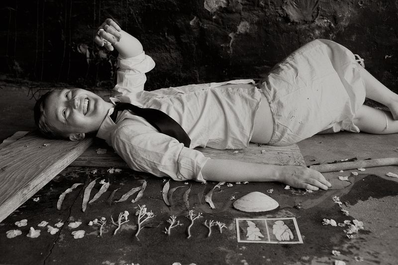 Black and white photo of a smiling child in a shirt and tie lying on a board with flowers, leaves, a shell, and pictures displayed.