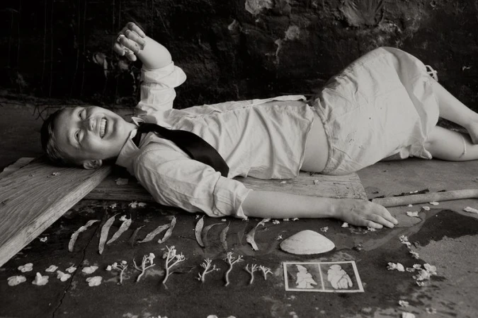 Black and white photo of a smiling child in a shirt and tie lying on a board with flowers, leaves, a shell, and pictures displayed.