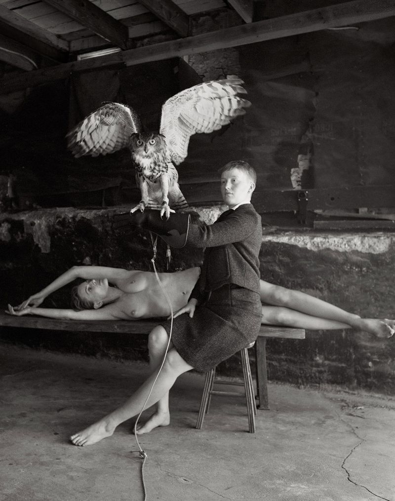 A person sits on a stool holding an owl, while a nude figure lies on a bench, captured in a black and white indoor scene.