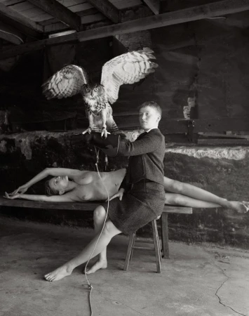 A person sits on a stool holding an owl, while a nude figure lies on a bench, captured in a black and white indoor scene.