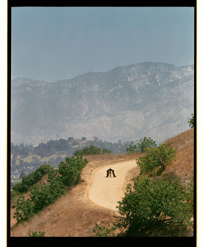 Two people stand facing each other on a dirt road surrounded by lush greenery with a mountain range visible in the background under a clear sky.