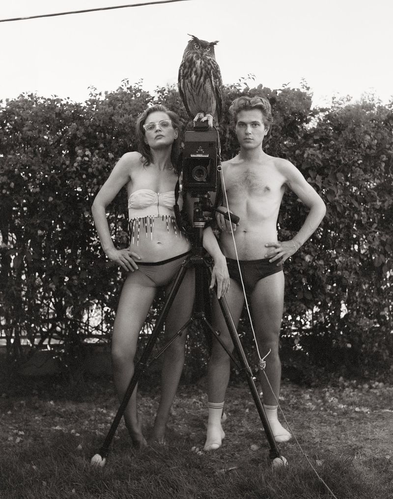 Monochrome shot featuring a man and a woman standing beside a large-format camera on a tripod, topped with an owl, set in a yard.