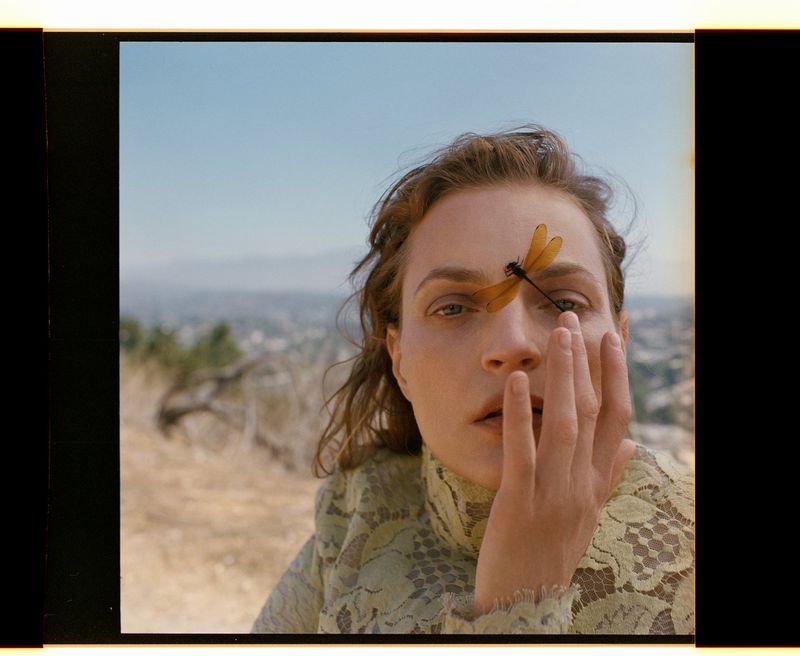 Close-up of woman with dragonfly on forehead, wearing a lace top, set against an outdoor landscape with a clear sky.