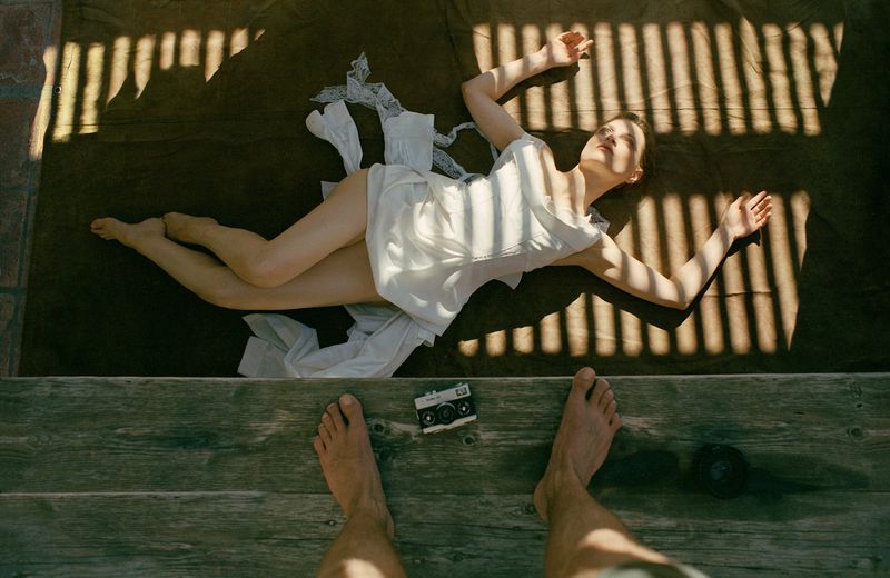 A woman in a white dress lays on a brown surface with shadows, with a vintage camera and a person's feet visible in the foreground.