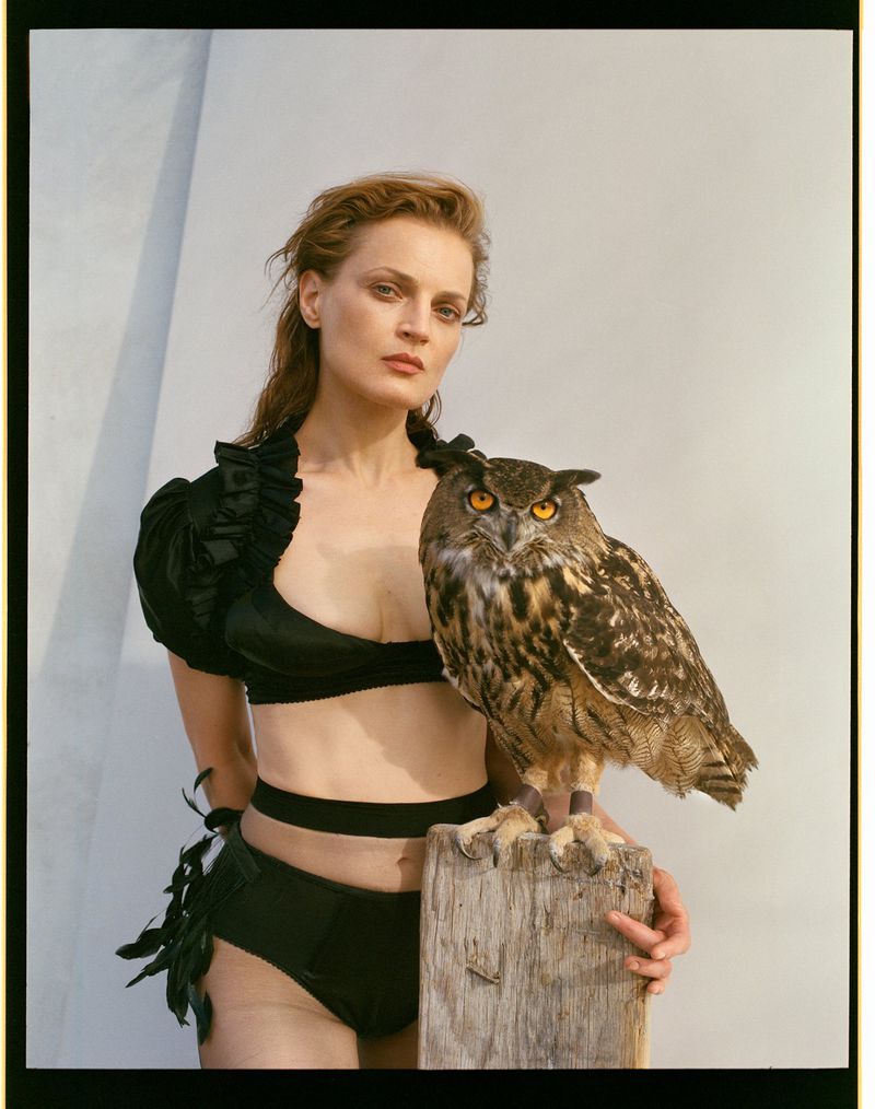 Adult woman in dark lingerie and puff-sleeve blouse poses with a European Eagle-Owl perched on a wooden post in a studio setting.