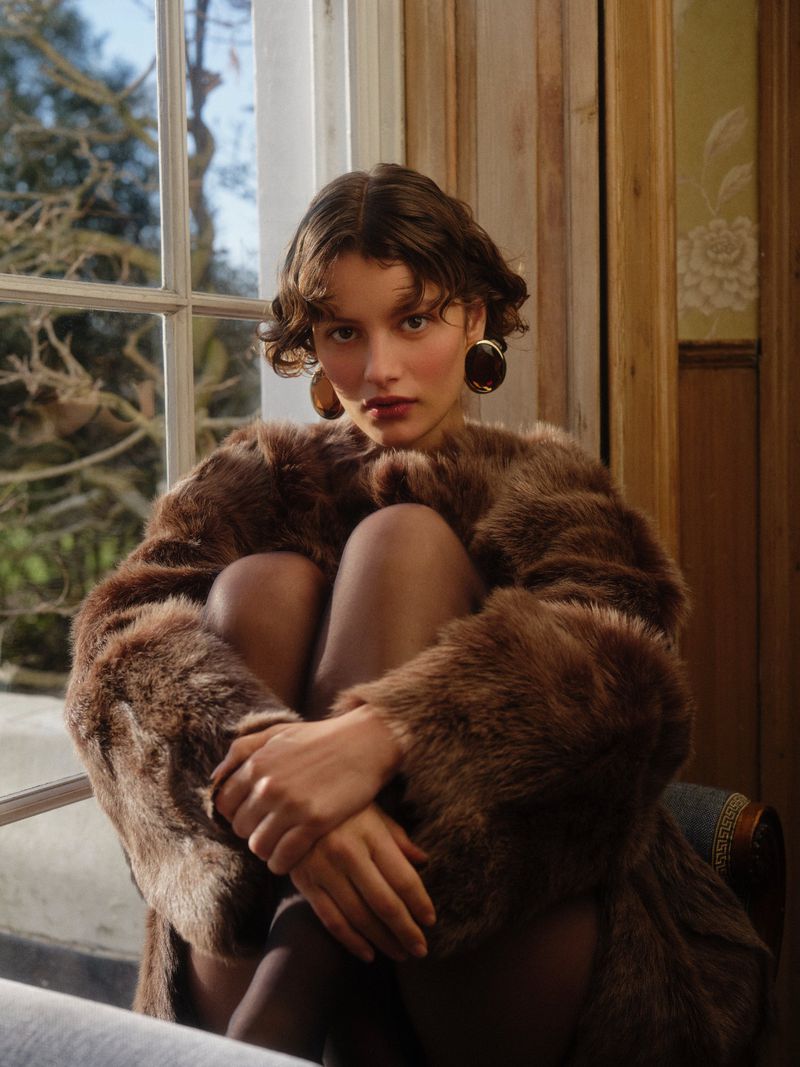 Portrait of woman in fur coat posing indoors near window. Adult female with wavy brown hair and dark eye makeup wears large earrings.