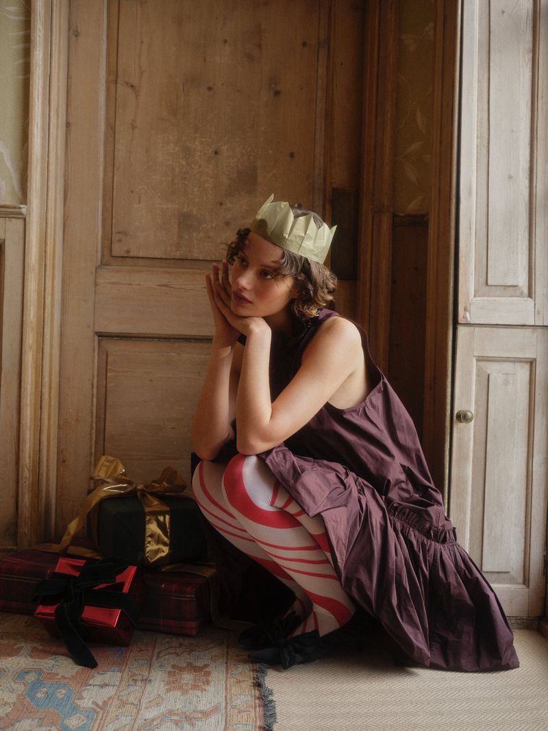 Young adult woman with paper crown poses with gifts in an indoor setting featuring a rustic door and vintage-style rug.