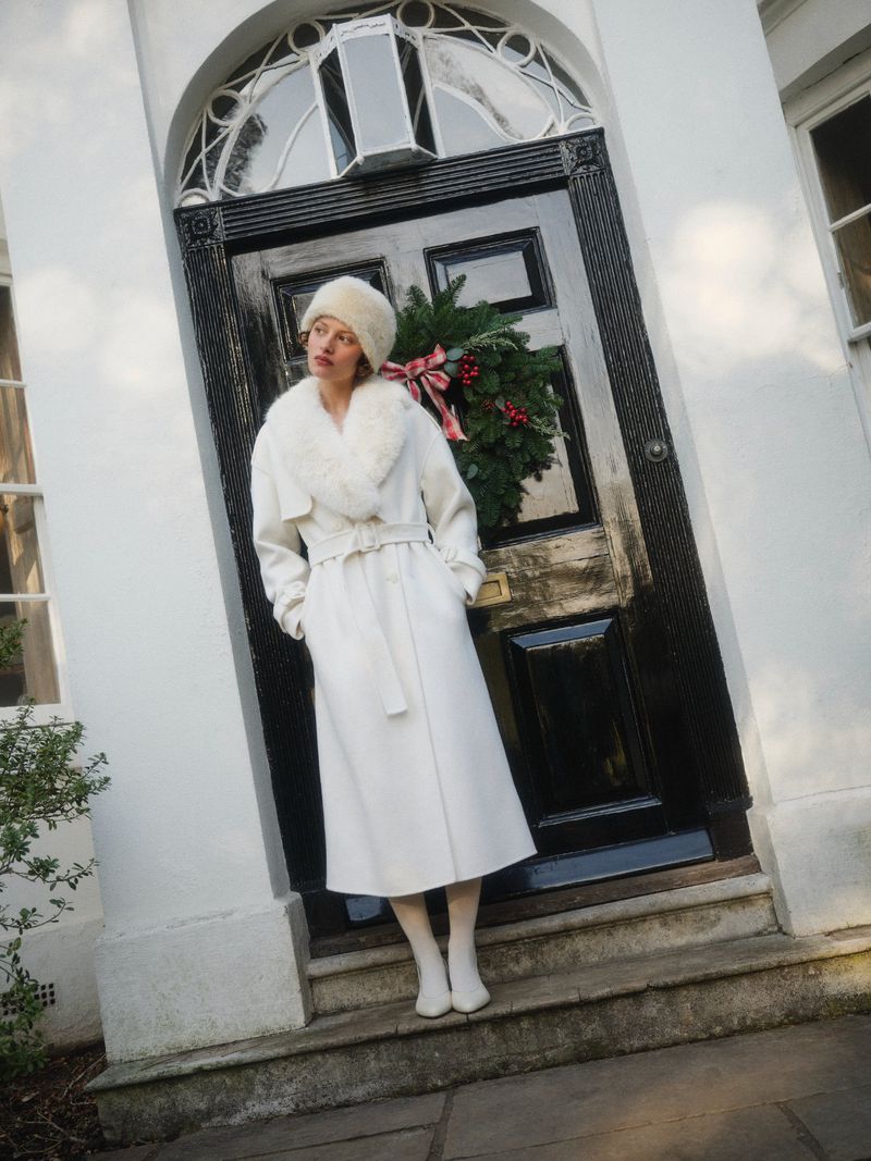 Woman in white coat and fur hat stands on steps near black door with wreath. Fashion shot in front of white building facade.