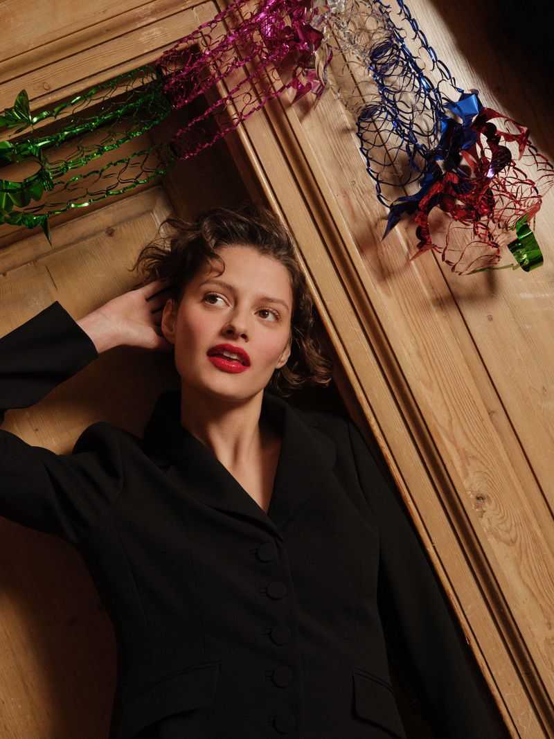Portrait of a young adult woman in a black blazer with bright red lipstick, reclining against a wooden paneled wall decorated with tinsel.
