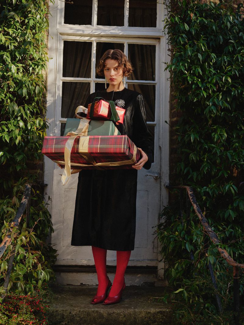Elegant woman in a black dress and red tights stands outside a white door with ivy, holding wrapped Christmas gifts with ribbons.