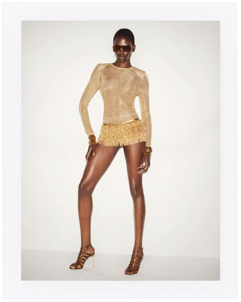 Glamorous African-American female model in a gold fringe mini skirt, matching top, bracelet cuffs and strappy heels posing in studio.