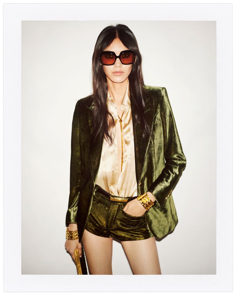 Brunette woman poses in olive-green velvet blazer and shorts, accessorized with gold bracelets and belt, wearing sunglasses against a white background.