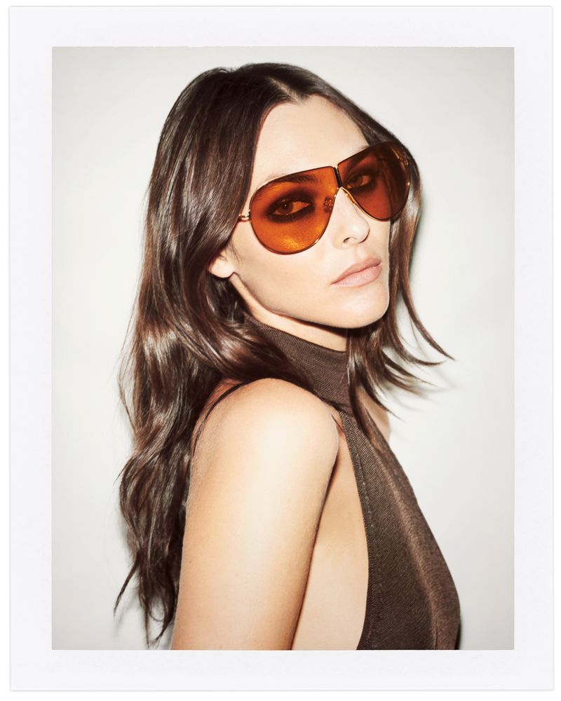 A brunette woman poses indoors wearing orange-tinted aviator sunglasses and a brown, high-neck dress against a light background.