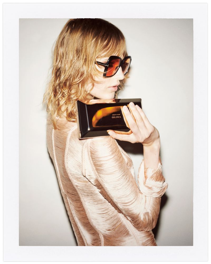 A fair-skinned woman with wavy blonde hair poses in a fringed top and sunglasses, holding a perfume box against a neutral backdrop.