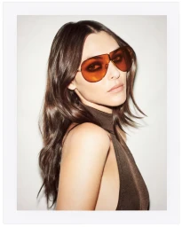 Model wearing amber-tinted aviator sunglasses and a brown sleeveless top against a white seamless background.