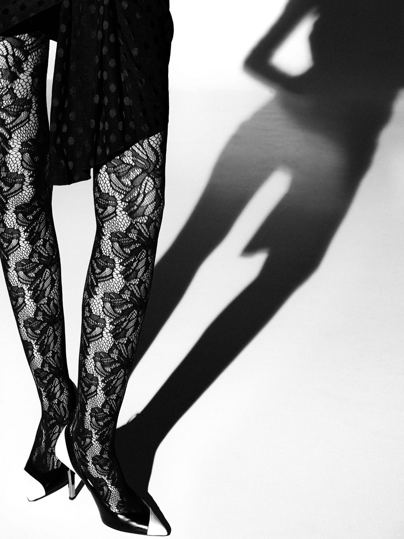 Close-up of legs in black lace tights and cap-toe pumps, casting a human shadow on a white background, creating a fashion-forward and artistic shot.