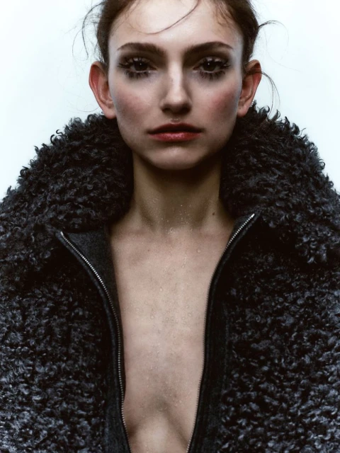 Close-up studio portrait of a young adult woman wearing a textured grey coat, focusing on her face and neutral expression, with clean background.