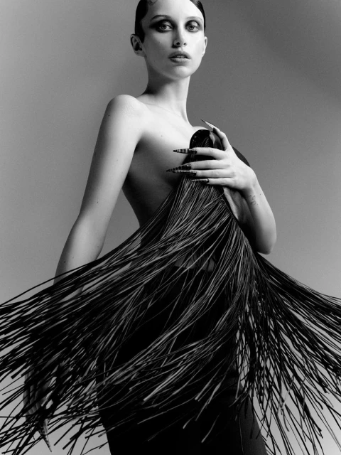 Striking black-and-white portrait of a young woman with dark hair, dramatic eye makeup, and long nails, wearing a distinctive fringe dress.