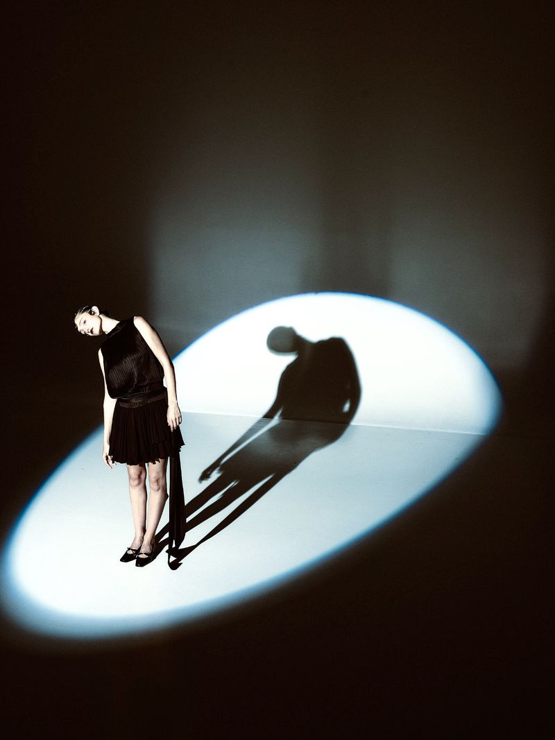 A woman in a black dress stands in a spotlight, casting a long shadow on a dark background, evoking a sense of mystery and performance.