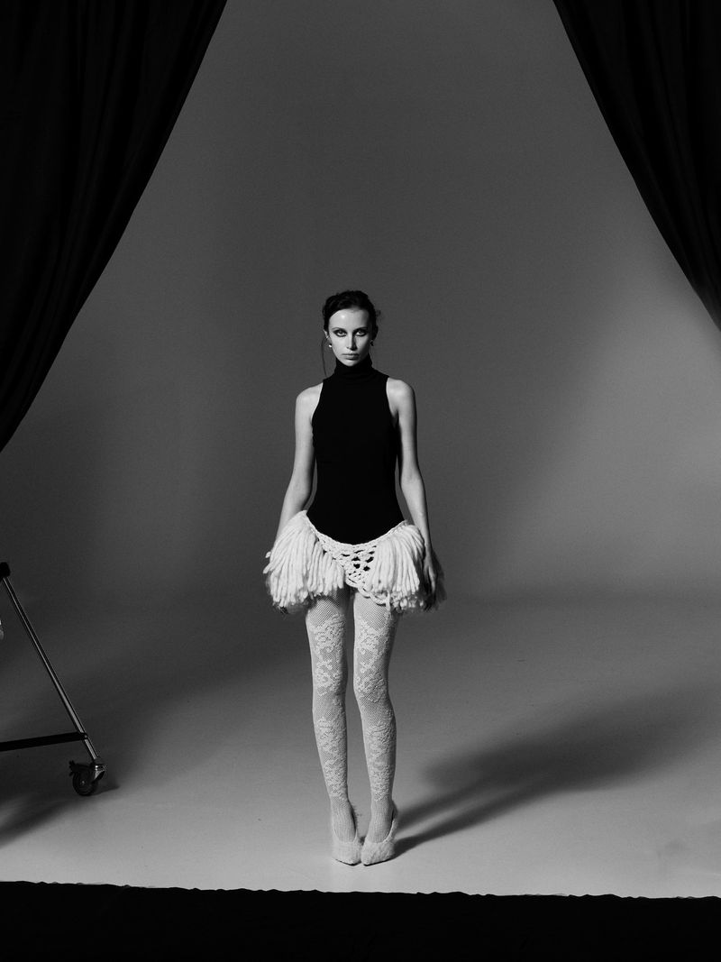 Fashion model poses in a black bodysuit, white lace tights, and matching skirt with fringe in a photo studio with dark drapes.