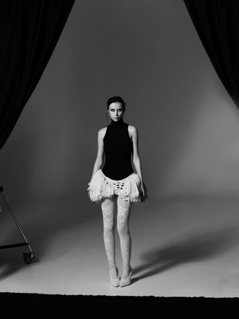 Fashion model poses in a black bodysuit, white lace tights, and matching skirt with fringe in a photo studio with dark drapes.