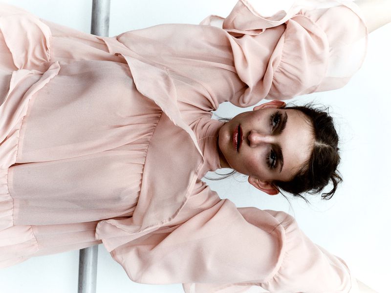 Portrait of a woman in a light pink dress with ruffled sleeves against a plain white background in a vertical, eye-level shot.