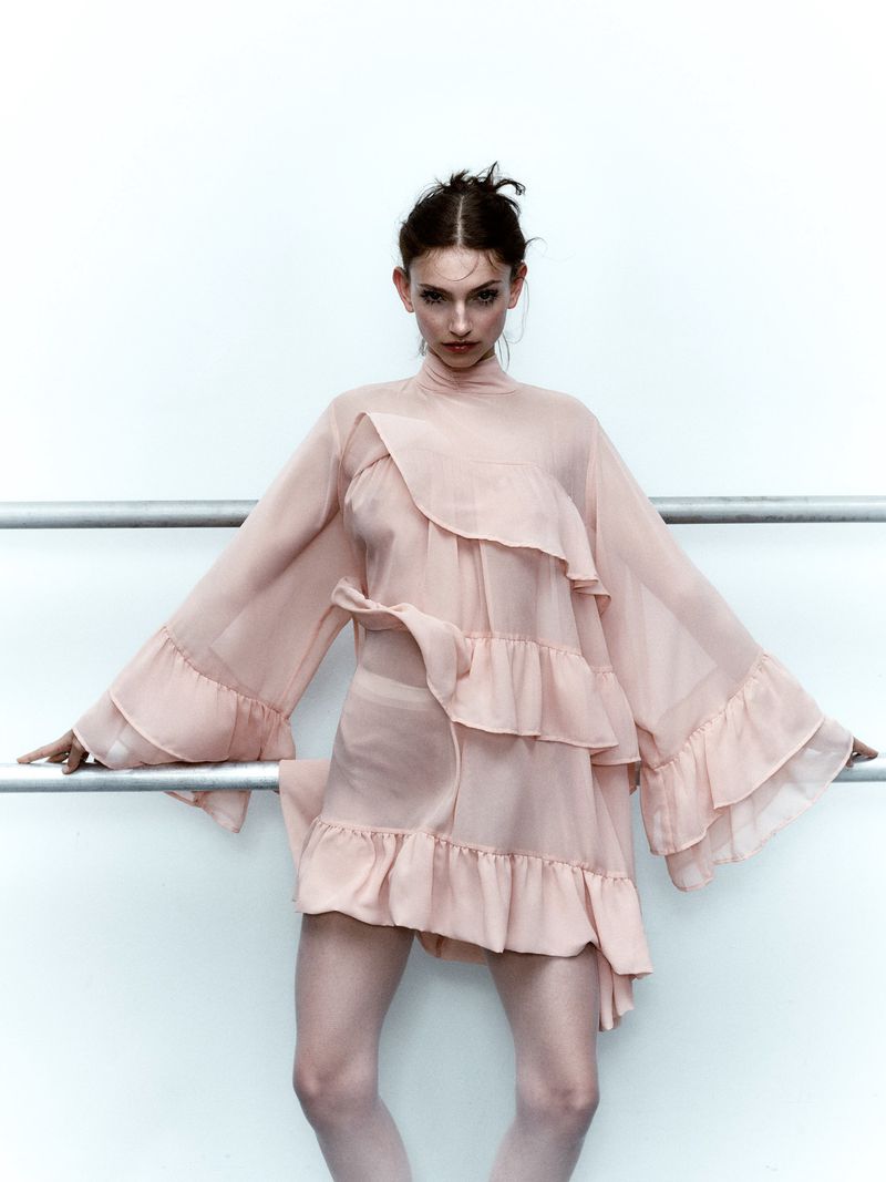 A stylish adult woman with neutral expression poses in a light pink dress with ruffles against a white background with metal bars.