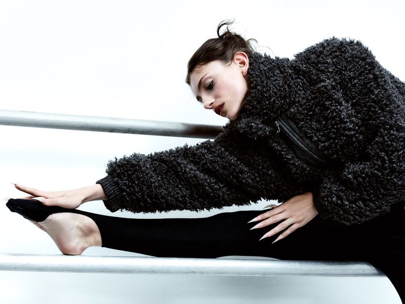 Adult female stretching in a studio, wearing a black textured jacket and leggings, reaching toward her outstretched leg in a modern dance pose.