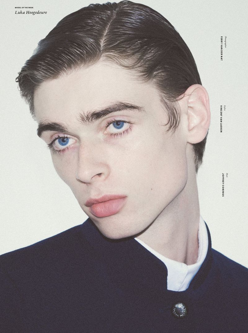 Portrait of a fair-skinned male model with blue eyes wearing a dark-blue jacket and white collared shirt against a white background.