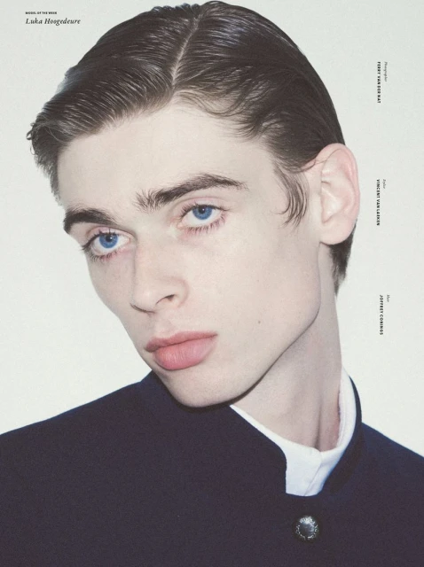 Portrait of a fair-skinned male model with blue eyes wearing a dark-blue jacket and white collared shirt against a white background.
