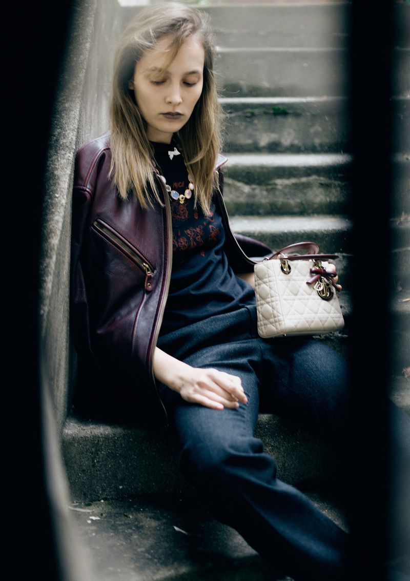 Fashionable adult woman poses on concrete steps wearing a leather jacket, dark top, jeans, and carrying a cream-colored handbag outdoors.