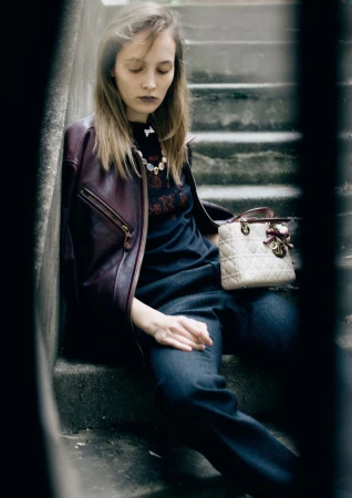 Fashionable adult woman poses on concrete steps wearing a leather jacket, dark top, jeans, and carrying a cream-colored handbag outdoors.