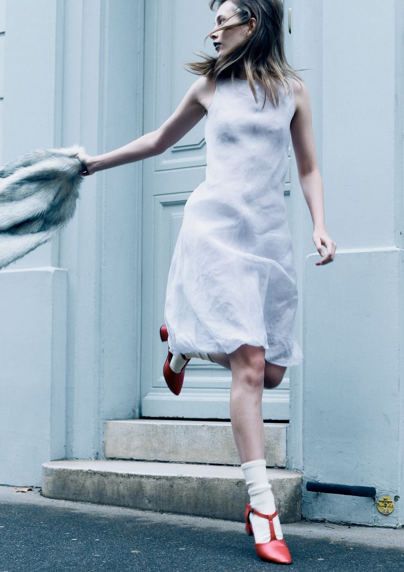 Woman in white dress and red heels running past a light blue building, holding a fur stole. Fashion shot, outdoor setting, day.