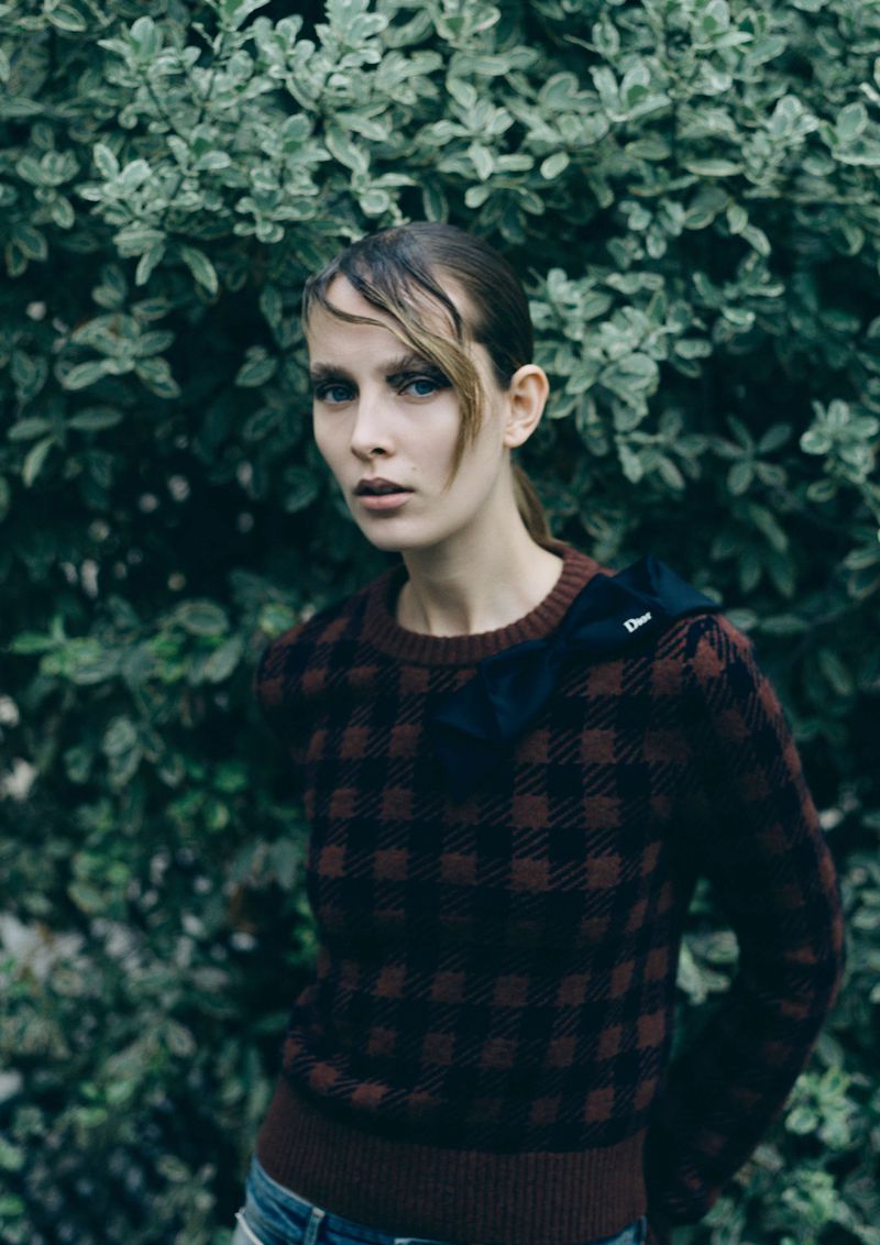 A stylish adult female model poses outdoors wearing a brown plaid sweater, with blurred green foliage filling the background, fashion editorial.