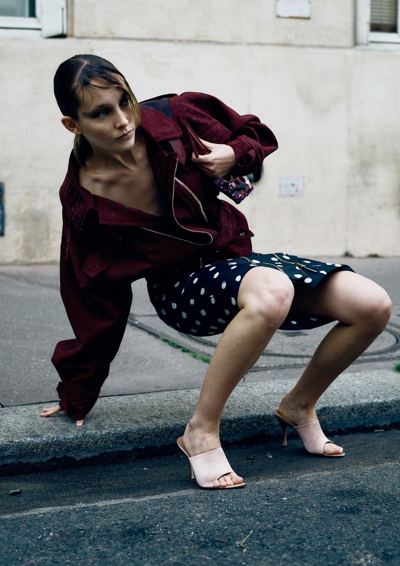 Fashion shot of a woman in a burgundy jacket and polka dot skirt posing on a city street next to a curb on a grey day.