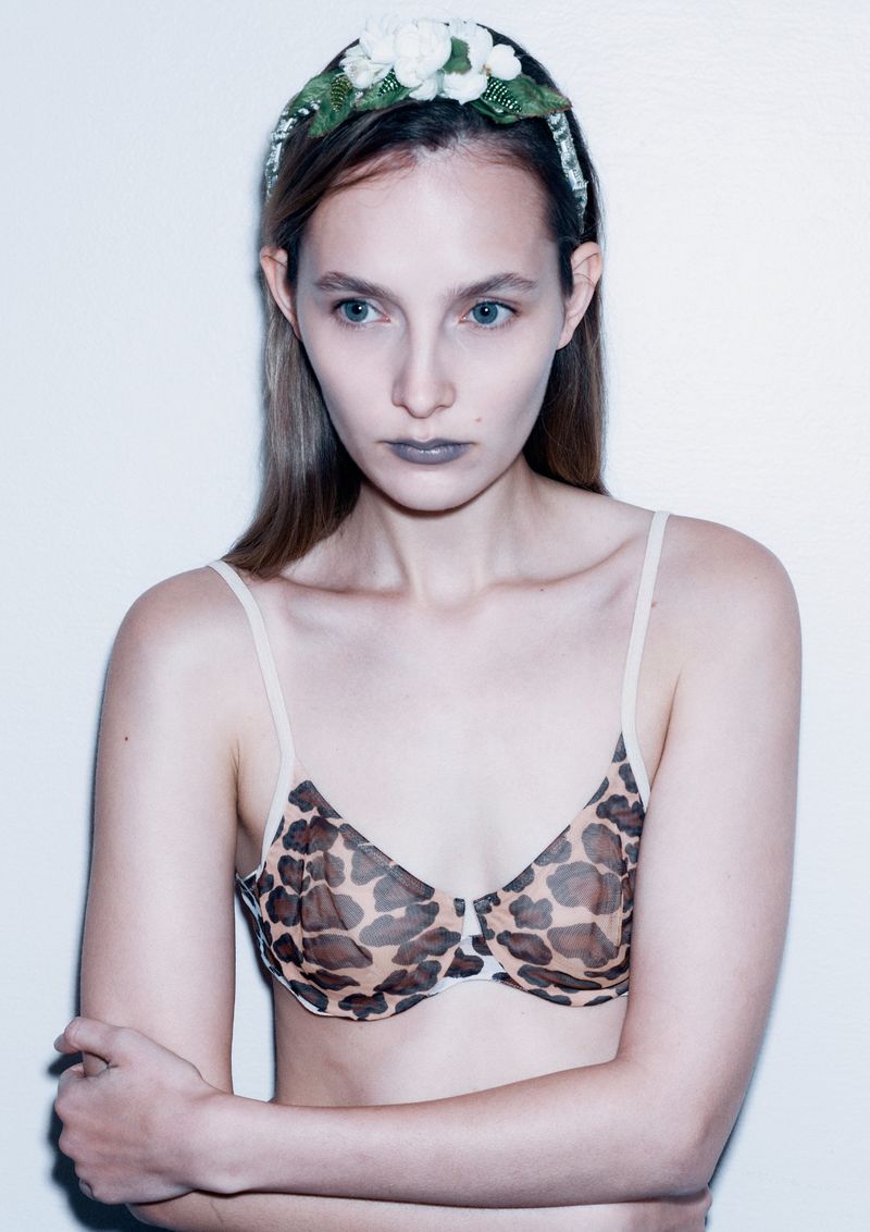 A fashionable woman with fair skin, wearing a flower crown and a leopard print bra, poses against a bright, neutral-toned background.