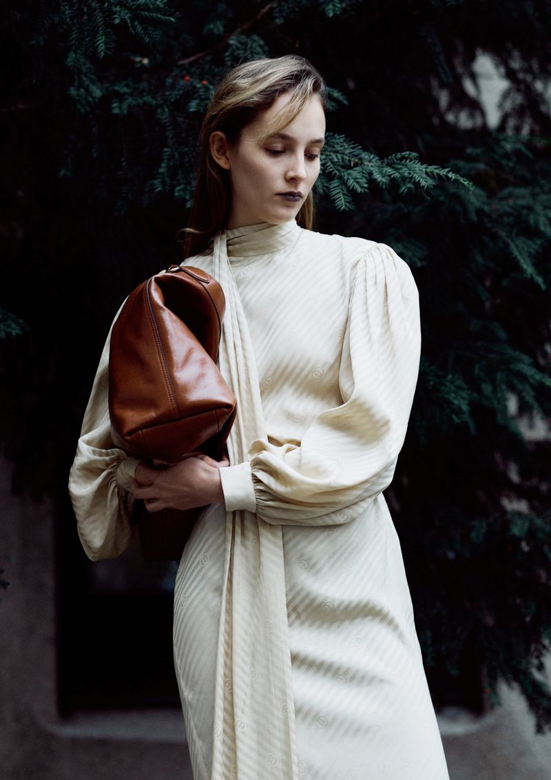 Woman in elegant dress holds leather purse in outdoor setting with natural lighting, posing against evergreen tree backdrop.