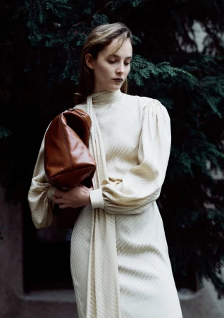 Woman in elegant dress holds leather purse in outdoor setting with natural lighting, posing against evergreen tree backdrop.
