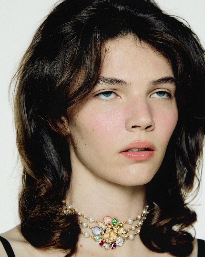 Close-up shot of a young woman with dark wavy hair and a jeweled necklace, looking up against a plain white backdrop.