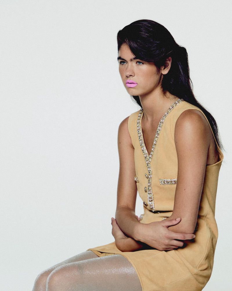 A young woman with long dark hair, wearing a light yellow dress and sitting with crossed arms, poses against a white backdrop.