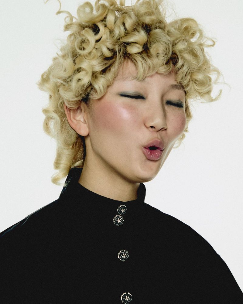 Portrait of an Asian woman with blonde curly hair, wearing a black top and pursing her lips, set against a clean white backdrop in a studio setting.