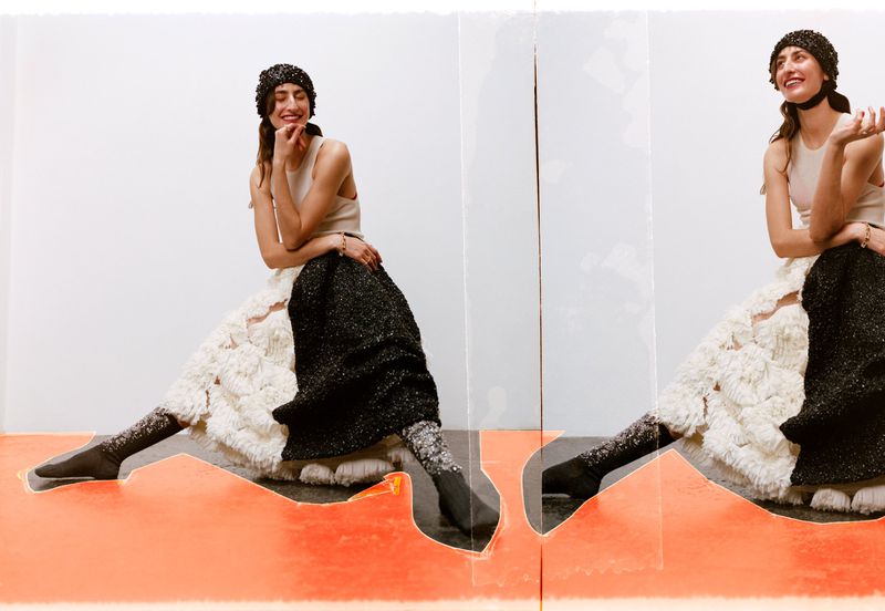 A cheerful model poses in a light-beige tank top and elaborate black-and-white skirt, set against a layered background of white and orange.