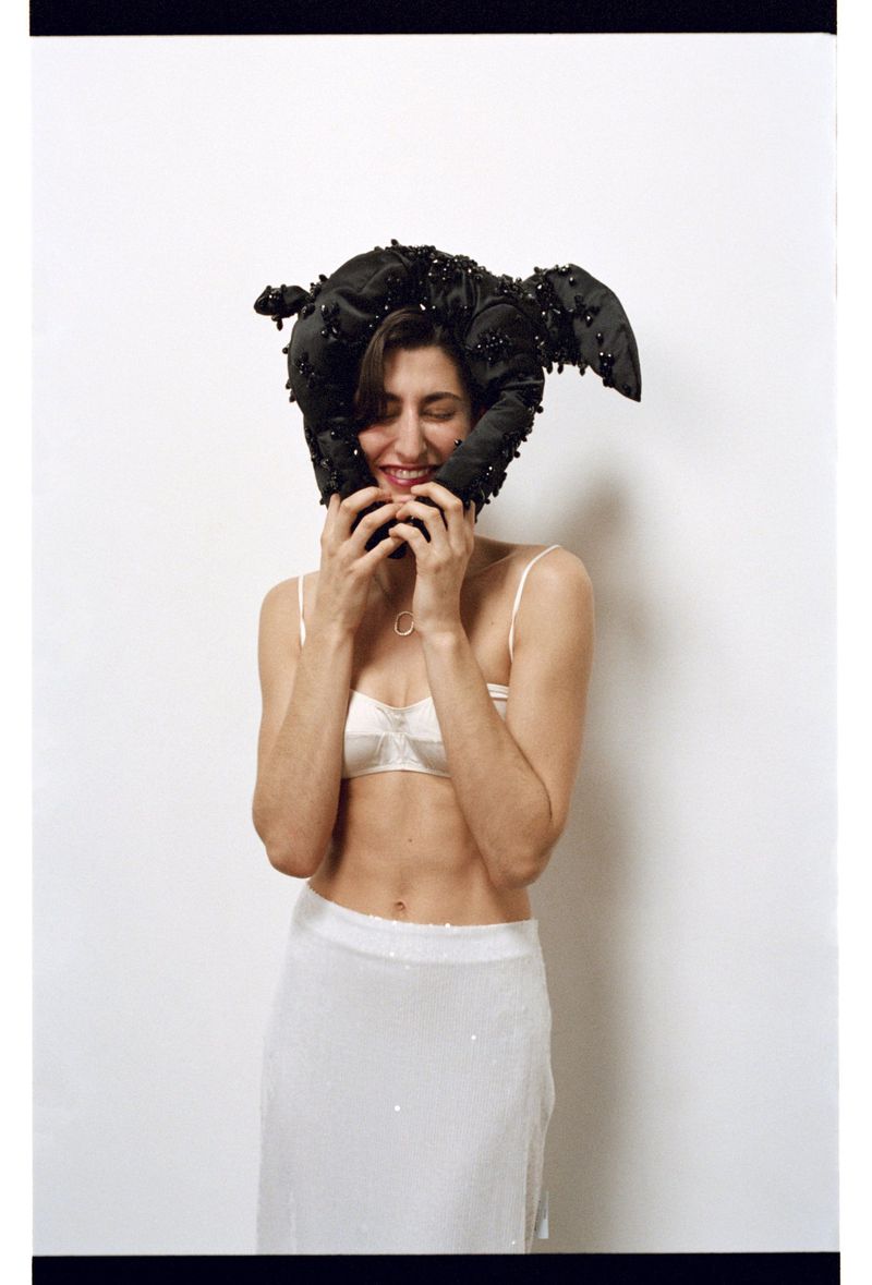 Smiling woman poses in front of a white wall with a black, embellished headpiece, a white lingerie top, and a long, light skirt.