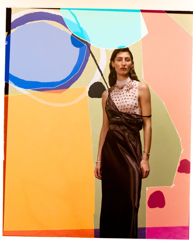 Elegant woman in a brown satin dress poses against an abstract geometric background with orange, blue, and other pastel colors in an indoor setting.