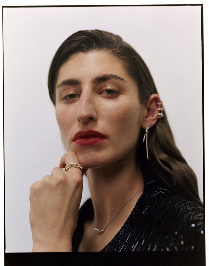 Close-up studio portrait of a woman with long brown hair wearing a black sequined jacket and gold jewelry against a white backdrop.
