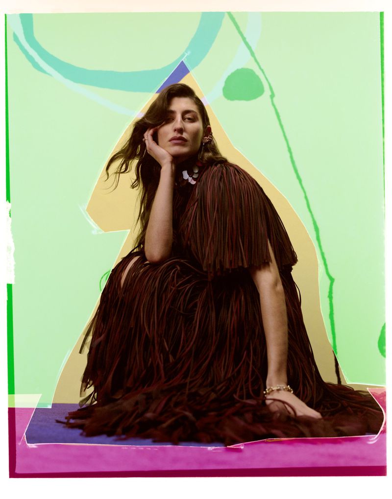 Brunette woman in a brown, fringed dress poses seated against an abstract background of light green, violet, and peach tones in a studio shot.