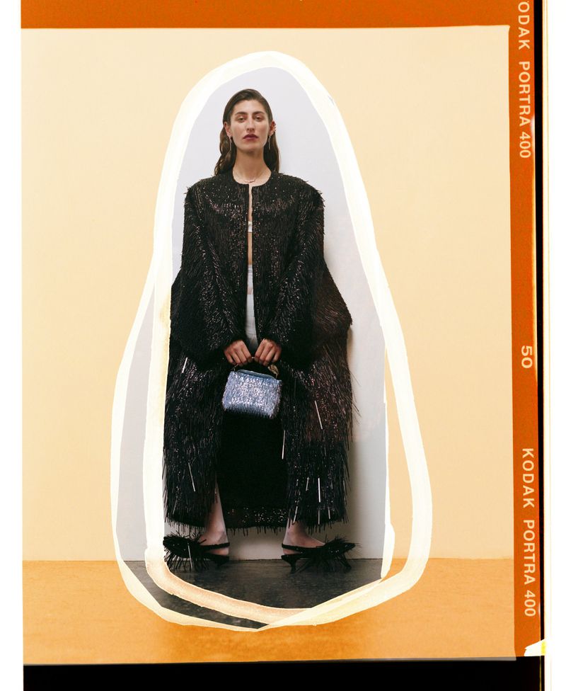 A woman sits in a long black coat and sparkly purse against a tan background in a studio setting; dark hair, neutral expression.