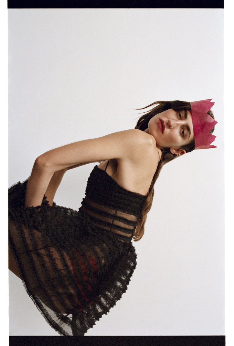 Woman wearing a black dress and a red paper crown, leaning backward on a white backdrop, studio lighting, medium shot, fashion editorial.