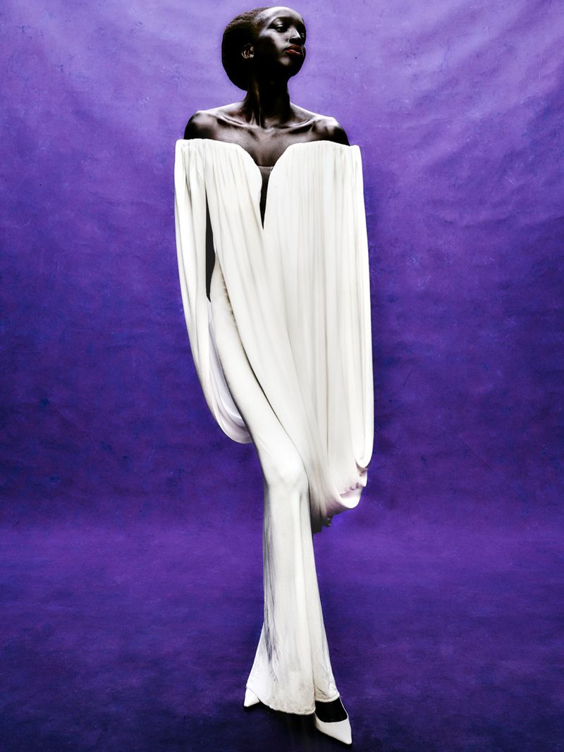 Woman poses wearing an off-the-shoulder white gown in a studio. Full shot against a dark purple backdrop with a shallow depth of field.