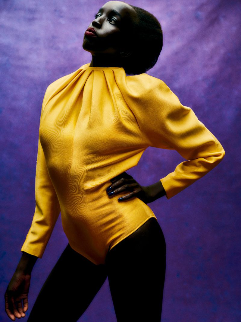 Fashion portrait of a Black woman in a yellow bodysuit against a purple backdrop, showcasing a modern and stylish look with clean lines.